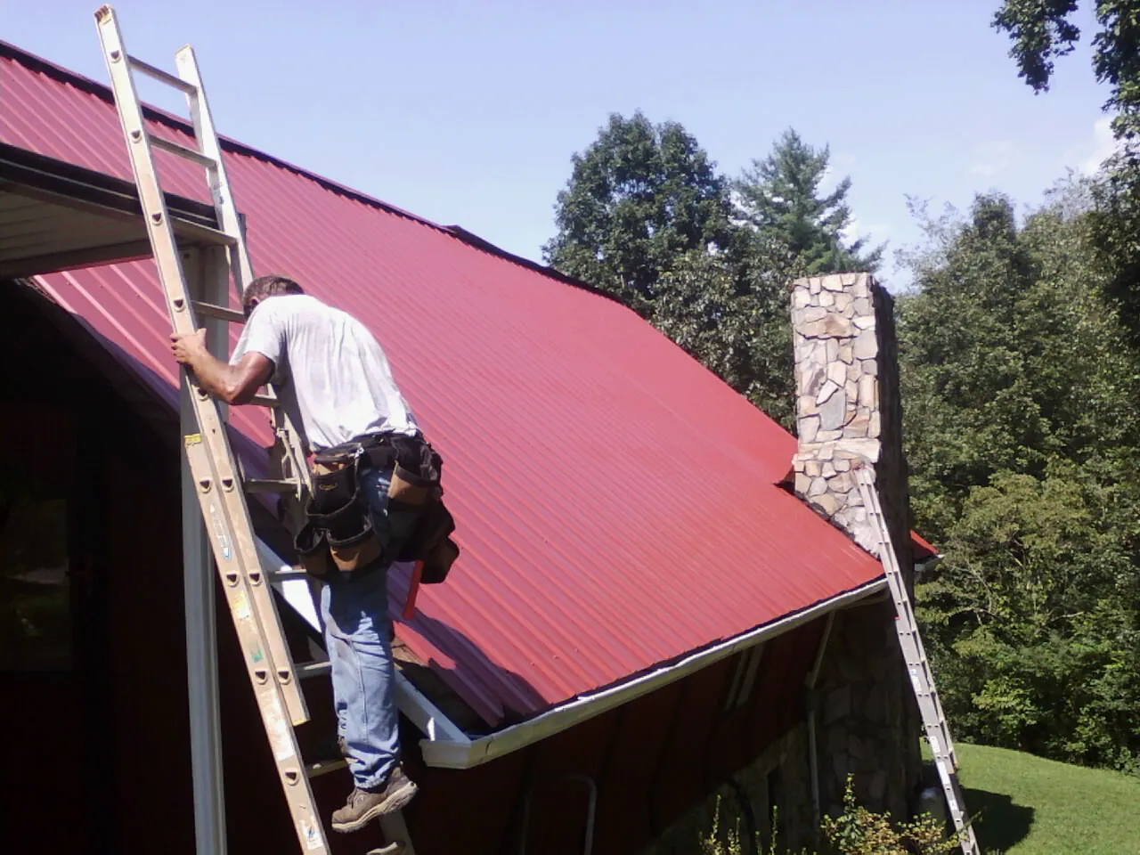 Roofer on a ladder beside a red metal roof and stone chimney — Soffit & Fascia Repair by Roofing Brothers in Monument